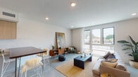 This contemporary single-floor residence showcases a thoughtfully designed open-concept layout that seamlessly connects the living, dining, and kitchen areas. Polished concrete floors run throughout, lending an industrial-modern aesthetic that ties the entire space together.

The living area is bright and welcoming, anchored by expansive floor-to-ceiling windows that flood the room with natural light and offer views of the surrounding neighborhood. Recessed lighting complements the natural light, while clean white walls provide a neutral backdrop ideal for art and decor.

The kitchen is well-appointed with flat-front cabinetry in a warm wood tone paired with light lower cabinets, white subway tile backsplash, stainless steel appliances including a gas range, microwave, dishwasher, and refrigerator, and a dark wood breakfast bar with bar seating — perfect for casual dining.

The bedroom is a calm, understated retreat with light hardwood-style flooring, soft neutral walls, and recessed lighting, offering a tranquil space for rest.

The bathroom features a frameless glass walk-in shower clad in white tile with a built-in niche, a floating wood-toned vanity with a white countertop, a wood-framed mirror, and polished concrete floors that echo the rest of the home.

Overall, the space presents a cohesive modern design with warm natural materials balanced against clean, minimalist finishes.