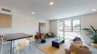 This contemporary single-floor residence showcases a thoughtfully designed open-concept layout that seamlessly connects the living, dining, and kitchen areas. Polished concrete floors run throughout, lending an industrial-modern aesthetic that ties the entire space together.

The living area is bright and welcoming, anchored by expansive floor-to-ceiling windows that flood the room with natural light and offer views of the surrounding neighborhood. Recessed lighting complements the natural light, while clean white walls provide a neutral backdrop ideal for art and decor.

The kitchen is well-appointed with flat-front cabinetry in a warm wood tone paired with light lower cabinets, white subway tile backsplash, stainless steel appliances including a gas range, microwave, dishwasher, and refrigerator, and a dark wood breakfast bar with bar seating — perfect for casual dining.

The bedroom is a calm, understated retreat with light hardwood-style flooring, soft neutral walls, and recessed lighting, offering a tranquil space for rest.

The bathroom features a frameless glass walk-in shower clad in white tile with a built-in niche, a floating wood-toned vanity with a white countertop, a wood-framed mirror, and polished concrete floors that echo the rest of the home.

Overall, the space presents a cohesive modern design with warm natural materials balanced against clean, minimalist finishes.