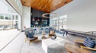 Located at 5580 Farrington Rd in Durham, NC, this generously sized amenity facility offers a seamlessly connected indoor-outdoor experience designed for relaxation and socializing. The interior clubroom is an architectural standout, boasting soaring double-height ceilings clad in warm, rich wood paneling that contrast beautifully with the crisp white walls and large-format tile flooring. A dramatic circular pendant light anchors the lounge area, complemented by bold red pendant lights that hang over a sleek, white quartz wet bar with dark cabinetry and patterned tile accents. Bar-height seating lines the counter, while a curated lounge area features stylish woven chairs and a geometric area rug. A shuffleboard table adds a recreational focal point to the space. Expansive floor-to-ceiling windows and wide bi-folding glass doors flood the interior with natural light and open directly onto the outdoor pool deck, creating a seamless indoor-outdoor connection. Outside, the large resort-style swimming pool is the centerpiece, surrounded by an expansive patio furnished with lounge chairs and shade umbrellas. The pool deck offers direct views of the multi-story residential building, set against a backdrop of mature trees, making this amenity space both functional and visually impressive.