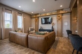 Located at 5060 City Station Dr in Collegedale, Tennessee, this single-floor community clubhouse offers a thoughtfully designed gathering space that blends modern aesthetics with warm, residential comfort. The interior is anchored by a sophisticated lounge and media area, where warm wood-paneled accent walls frame a wall-mounted flat-screen TV above a sleek dark console. Built-in open shelving runs overhead, lined with books and decorative accessories, while stylish sconce lighting adds a refined ambiance. Seating includes a generous cognac leather sectional, accent chairs, and a fluted round coffee table, creating an inviting space for relaxation and social interaction. Adjacent to the lounge, a compact co-working nook features built-in cabinetry with a quartz countertop, a geometric-patterned tile feature wall in muted teal tones, and a dedicated workspace with a printer station. A separate gathering room showcases soaring ceilings, floor-to-ceiling windows that flood the space with natural light, a striking woven pendant light fixture, a long communal bar-height table, and an eclectic mix of seating including plaid wingback chairs and green velvet ottomans. Light travertine-style flooring flows consistently throughout, unifying the space with a clean, contemporary feel.