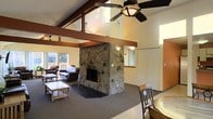 This two-floor space blends rustic character with comfortable living across a well-designed layout. The main living area immediately draws attention with its soaring vaulted ceilings, accented by rich, dark-stained exposed wood beams that add structural elegance and a classic lodge aesthetic. A ceiling fan is centrally mounted, promoting airflow throughout the generous open space.

The undeniable focal point of the main level is a floor-to-ceiling stone fireplace, constructed from natural stacked rock. Its substantial presence anchors the living room and adds a rugged, cabin-inspired warmth to the interior. The surrounding seating area is furnished with leather upholstered sofas and a wooden coffee table, creating an inviting space for gathering.

Natural light filters in through multiple windows, including a view toward what appears to be an outdoor deck, enhancing the connection between interior and exterior environments. The flooring in the main living area is wall-to-wall carpet, contributing to the cozy, relaxed atmosphere.

A dining area is visible adjacent to the living space, furnished with wooden chairs, suggesting a seamless flow between living and dining zones. Additional rooms are accessible through interior doorways, and the second floor offers further living accommodations. Overall, this space exudes a warm, lodge-style ambiance well-suited for relaxed, comfortable living.