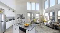 This single-floor space is defined by its dramatic double-height ceilings and an open-concept layout that connects the kitchen and living area in one cohesive environment. Oversized windows positioned high on the walls flood the interior with natural light, enhancing the airy, expansive atmosphere throughout.

The kitchen is anchored by a central island with a sleek grey countertop, double stainless steel sink, and integrated dishwasher. White cabinetry lines the perimeter, contributing to a clean, contemporary aesthetic. The island serves as both a functional workspace and a natural divider between the kitchen and the adjoining living space.

The living area is generously proportioned, featuring large glass doors that frame outdoor views and invite additional natural light. A patterned area rug grounds the seating arrangement, which includes a grey sofa accented with yellow cushions, adding a vibrant pop of color against the predominantly neutral palette. A glass-top coffee table and coordinated side furniture complete the arrangement.

Throughout the space, the white walls, light flooring, and carefully selected accents create a modern, welcoming ambiance that feels both functional and stylish.