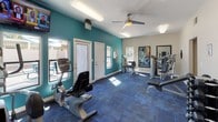 This fitness center offers a practical and energizing environment for exercise and wellness. The space is defined by its bold teal accent walls, which lend a vibrant, motivating atmosphere, complemented by blue modular carpet flooring that provides cushioning underfoot throughout the workout area. Natural light streams in through multiple windows, creating a bright and welcoming environment, while a ceiling fan ensures comfortable air circulation during workouts. The facility is equipped with a variety of cardio machines, including a recumbent bike and an elliptical trainer, as well as strength training equipment and a weight bench, catering to a range of fitness routines. Full-length mirrors mounted along one wall allow users to monitor their form and also enhance the sense of space within the room. Overhead fluorescent lighting supplements the natural light, ensuring the space remains well-lit at all hours. A clearly marked exit door and a frosted glass entry door provide convenient access points. The overall layout is open and functional, with equipment thoughtfully arranged to maximize usable floor space and allow for safe, comfortable movement between machines.