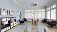 This single-story after school care space is designed to provide a comfortable and functional environment for students. The main room is generously proportioned, featuring light wood-look flooring, crisp white crown molding, and soft blue-gray accent walls that create a calm, inviting atmosphere. A large bank of white-framed windows lines one wall, flooding the space with abundant natural light and offering views of surrounding greenery. A ceiling fan with a modern profile enhances air circulation throughout the room. The space is thoughtfully arranged with multiple seating zones to accommodate a variety of activities. A dining-style table with vibrant orange chairs serves as a central gathering spot for group work or snacks, while a pair of sleek black swivel chairs and a small side table create a more relaxed lounge area. A low bookshelf with organized storage bins lines one wall alongside a curated display of framed educational artwork, adding character and visual interest. The open layout allows for easy supervision and flexible use, making it well-suited for homework, group projects, or quiet downtime.