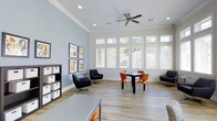 This single-story after school care space is designed to provide a comfortable and functional environment for students. The main room is generously proportioned, featuring light wood-look flooring, crisp white crown molding, and soft blue-gray accent walls that create a calm, inviting atmosphere. A large bank of white-framed windows lines one wall, flooding the space with abundant natural light and offering views of surrounding greenery. A ceiling fan with a modern profile enhances air circulation throughout the room. The space is thoughtfully arranged with multiple seating zones to accommodate a variety of activities. A dining-style table with vibrant orange chairs serves as a central gathering spot for group work or snacks, while a pair of sleek black swivel chairs and a small side table create a more relaxed lounge area. A low bookshelf with organized storage bins lines one wall alongside a curated display of framed educational artwork, adding character and visual interest. The open layout allows for easy supervision and flexible use, making it well-suited for homework, group projects, or quiet downtime.