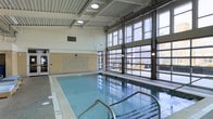This indoor aquatic facility is designed to provide a bright, welcoming environment for swimmers and leisure users alike. The space is anchored by a generously sized rectangular swimming pool with clearly marked lane depths, finished with clean white pool decking along its perimeter. Stainless steel entry rails offer convenient pool access at multiple points.

A separate spa or hydrotherapy pool is situated adjacent to the main pool, providing a dedicated relaxation area. The two bodies of water are thoughtfully positioned within the same open room, maximizing the usability of the space.

The architecture draws attention with its soaring ceiling fitted with a white ribbed panel system and recessed lighting that evenly illuminates the interior. A striking wall of large, industrial-style glass garage doors spans one side of the room from floor to ceiling, flooding the space with abundant natural light and offering views of the surrounding exterior buildings. This feature also allows the space to feel open and connected to the outdoors.

A decorative blue mosaic tile accent wall adds a subtle design detail, complementing the aquatic setting. The overall ambiance is clean, functional, and modern, suited for both recreational and fitness-oriented use.