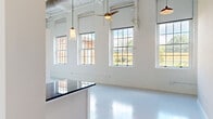 Located at 128 Mill St in Clayton, NC, this single-floor industrial loft conversion masterfully balances historic charm with contemporary design. The open main living area is a standout, showcasing whitewashed exposed brick walls, soaring ceilings with visible ductwork and piping, and a row of large multi-pane windows that flood the space with natural light and offer views of the surrounding streetscape. Ceiling fans and pendant lighting complement the loft's airy, industrial aesthetic, while polished concrete-style flooring runs seamlessly throughout. The well-appointed kitchen features an abundance of white cabinetry, dark granite countertops, a white subway tile backsplash, and a full suite of stainless steel appliances, including a refrigerator, range, microwave, and dishwasher. Track and pendant lighting add warmth to this functional space. The bedroom area benefits from tall ceilings and its own large window, bringing in natural light, and is accompanied by a dedicated closet with built-in shelving. The bathroom offers a clean, modern finish with white cabinetry, a dark-framed mirror, dark countertops, and a glass-enclosed tiled shower. Throughout, the seamless polished floors tie the entire unit together in a cohesive, modern aesthetic.