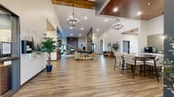 Located at 1920 Old Main St S in Cambridge, Minnesota, this single-floor community clubhouse — known as The Lodge — offers a generously sized, open-concept gathering space designed with a refined, nature-inspired aesthetic. The interior is anchored by dramatic vaulted ceilings with exposed wood beam accents, two elegant chandelier-style pendant light fixtures, and rich wood-look luxury vinyl plank flooring that flows throughout the entire space.

At the heart of the room, a floor-to-ceiling natural stone fireplace serves as a commanding focal point, surrounded by comfortable lounge seating and layered area rugs that create an inviting conversation area. Adjacent to this is a dedicated media lounge with a large wall-mounted television, a stylish storage console, and natural light streaming in through slatted window blinds.

A bar-height dining counter with seating and a nearby kitchenette area rounds out the social amenities, complemented by a branded "Lodge" signage wall that reinforces the community identity. Decorative indoor trees and curated artwork lend warmth and life to the neutral white and teal accent color palette.

The entryway corridor provides a welcoming transition into the space, with additional access doors, framed nature-inspired artwork, and a console table with organic décor. Outside, the building features stone-accented siding and resident balconies with ample parking and outdoor grilling stations nearby.