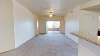 This single-floor space presents a straightforward and functional layout centered around a spacious main living area. The room is finished with neutral-toned wall-to-wall carpeting that extends throughout, contributing to a quiet and comfortable atmosphere. Smooth, light-colored walls reflect natural light effectively, keeping the interior feeling bright and airy.

A ceiling fan with an integrated light fixture is centrally mounted overhead, providing both ventilation and ambient illumination. A sliding glass door dominates the far wall, serving as the primary source of natural light and offering a direct view to an outdoor balcony area, where a railing and exterior greenery are visible. This connection to the outdoors enhances the sense of openness within the living space.

To one side, a partial countertop with a stone or granite-style finish is visible, suggesting a transition point between the living area and an adjacent kitchen or bar space. A secondary doorway is also visible near the far wall, indicating access to additional rooms or a hallway. The overall design is clean and understated, making it a versatile and adaptable living environment.