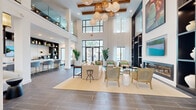 This elegantly designed communal lounge space makes a striking first impression with its soaring double-height ceilings, exposed wood beam accents, and floor-to-ceiling windows that flood the interior with natural light while offering views of lush tropical palms and a pool beyond. The open-concept layout is anchored by a generously sized seating area furnished with neutral-toned sofas, rattan accent chairs, and a glass coffee table atop a woven area rug, all centered around a sleek, built-in linear fireplace framed by a dramatic dark shelving unit displaying curated ceramic décor and vibrant botanical artwork. An upper-level mezzanine with glass railings overlooks the main living area, adding to the sense of grandeur. A dedicated wet bar and lounge counter runs along one side of the space, featuring light blue cabinetry, a white quartz countertop, open display shelving with white ceramic accents, a wall-mounted television, and stylish quilted blue bar stools with gold-toned legs. Dark wood-look tile flooring grounds the space throughout, complemented by a patterned tile entry area. A cluster of globe and rattan pendant lights serves as a decorative focal point overhead, tying together the space's coastal-modern aesthetic.
