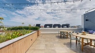 Located in the heart of Los Angeles along W Olympic Boulevard, this rooftop terrace offers a well-appointed open-air amenity space designed for both relaxation and social gatherings. The deck is divided into distinct functional zones, each finished with high-quality materials that blend modern and natural aesthetics seamlessly.

One section features warm wood-tone tile flooring paired with slatted wood-paneled planter boxes filled with lush greenery and flowering plants, creating a garden-like atmosphere. This area is outfitted with multiple dining tables dressed in blue gingham linens, white chairs, and blue outdoor rugs, all set beneath overhead string lights that add a festive ambiance. Adjacent to this zone, a built-in outdoor kitchen provides two stainless steel grills, prep surfaces, and ample counter space, making it ideal for catered events or communal cooking.

A separate lounge area is finished with large-format neutral stone tiles, featuring a round teak dining table with sage green chairs and a comfortable sofa seating arrangement against a striking dark blue textured stone accent wall. Architectural black sculptural elements line the parapet, adding a bold design touch.

Floor-to-ceiling glass windows and sliding glass doors connect the terrace to an interior space, allowing natural light to flow freely. The open sky above and panoramic views of the surrounding Los Angeles skyline complete this versatile rooftop destination.