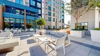 Located at 111 Green St SE in Gainesville, Georgia, this outdoor amenity space is a thoughtfully designed courtyard nestled within a modern, multi-story building complex. The architecture features a clean contemporary aesthetic, with light-toned paneled facades, large glass windows, and bold blue accent elements that frame the outdoor area on multiple sides.

The courtyard is organized into several distinct zones. A spacious open lawn area, finished with lush artificial turf, serves as a central gathering point and is furnished with white Adirondack chairs arranged in a social circle. Flanking the lawn, a wide paved terrace provides ample room for outdoor seating, featuring sleek white lounge furniture, accent tables, and umbrella-shaded dining sets ideal for relaxing or socializing. String lights are strung overhead, adding ambiance during evening hours.

A dedicated putting green area offers a recreational amenity for golf enthusiasts, complete with multiple holes and well-maintained synthetic turf. Additional landscaping elements, including large planter boxes with ornamental trees and greenery, add natural warmth and visual interest throughout the space.

The courtyard benefits from abundant natural light and open sky exposure, creating a bright and inviting atmosphere that encourages outdoor activity and community engagement.