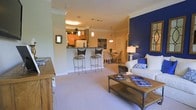 This single-story space is designed with an open-concept layout that connects the kitchen, dining, and living areas into one cohesive, flowing environment. The kitchen features warm wood cabinetry, a dark-topped breakfast bar with elegant wrought iron barstools, pendant lighting, and a white refrigerator, providing both function and style. Crown molding runs throughout, adding a polished architectural detail that elevates the overall aesthetic.

The dining area sits just beyond the kitchen and is furnished with a classic table and upholstered chairs beneath a hanging light fixture, creating an inviting space for meals and gatherings. The living area is anchored by a white sofa dressed with blue accent pillows, complemented by a rustic wood-and-metal coffee table that adds an industrial touch. A striking deep blue accent wall serves as a bold focal point, contrasting beautifully with the warm beige carpeting and cream-toned walls found throughout.

Natural-toned carpeting flows across the entire space, contributing to a warm and comfortable ambiance. The overall design balances classic architectural details with contemporary furnishing choices, creating a welcoming and well-appointed interior.