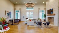 This single-floor community clubhouse is designed to offer residents a comprehensive range of amenities within a cohesive, modern aesthetic. The main entrance welcomes visitors through elegant double glass doors set beneath soaring ceilings, with a distinctive herringbone-patterned tile floor creating a polished first impression.

The heart of the facility is a grand lounge featuring dramatic double-ring chandelier lighting, warm hardwood-style flooring, and an abundance of natural light from large floor-to-ceiling windows. The space is furnished with plush sectional sofas, armchairs, and small groupings ideal for socializing or relaxing. An adjacent seating area offers a large flat-screen TV and additional lounge seating arranged on a patterned area rug.

The communal kitchen and dining area is outfitted with granite countertops, white cabinetry, subway tile backsplash, and a stainless steel sink. A long communal dining table with pendant lighting above provides ample space for group gatherings. A separate lounge nook with a TV and casual seating adjoins this area.

The fitness room includes treadmills, ceiling-mounted TV, and windows overlooking the exterior terrace. A professional leasing office area features multiple private consultation rooms and dedicated workstations. The space also includes a well-finished restroom with marble-topped vanity and decorative tile accents.