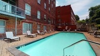 Located at 402 N 18th St in Richmond, Virginia, this single-floor common area showcases a compelling blend of historic industrial character and modern updates. The interior hallways are defined by exposed red brick walls, natural stone tile flooring, and original concrete structural columns that speak to the building's storied past as a repurposed industrial or warehouse structure. Flush-mounted ceiling fixtures illuminate the corridors, which stretch generously and provide clear sightlines from end to end, conveying a strong sense of depth and openness. Large glass doors and floor-to-ceiling windows allow natural light to filter into the entryway areas, connecting the interior to the surrounding outdoor spaces. Stairwell access is marked by a solid wood door set within the exposed brick, maintaining the building's raw aesthetic throughout. The highlight of the property is the expansive outdoor pool courtyard, framed on multiple sides by the building's striking multi-story red brick facade. The pool area features a generous sun deck with lounge chairs, a paved surround, and a shaded seating area, creating an inviting retreat. Together, the interplay of historic architectural materials and thoughtful amenities makes this a distinctive and functional space in the heart of Richmond.