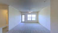 Located in Roanoke, TX, this single-story space features a spacious main living area designed with simplicity and functionality in mind. The room is finished with wall-to-wall carpet flooring in a neutral tone, contributing to a clean and understated aesthetic throughout. Smooth, light-colored walls are framed by subtle crown molding, adding a modest architectural detail that ties the space together.

Natural light plays a significant role in the ambiance of the living area. A large picture window positioned along one wall allows ample daylight to fill the room, while a glass-paneled exterior door nearby reinforces the bright, airy feel and provides direct access to the outdoors. The combination of these two openings ensures the space remains well-lit throughout the day.

Overhead, a ceiling fan with an integrated light fixture is centrally mounted, offering both air circulation and supplemental lighting. A standard HVAC vent is also visible in the ceiling, indicating a central climate control system. The overall layout is open and versatile, providing a blank canvas adaptable to a variety of furniture arrangements and uses.