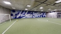 This indoor futsal facility offers a generously sized, single-level playing surface covered in green artificial turf, complete with clearly marked white boundary lines, a center circle, and goal boxes at each end. Two full-size portable goals are positioned at opposing ends of the pitch, making the space ready for competitive or casual play. The structural aesthetic is defined by exposed concrete walls and ceiling, lending an industrial character that is both raw and functional. Overhead, a grid of recessed and surface-mounted fluorescent panel lights ensures even, bright illumination across the entire playing area. One wall is dramatically transformed by a large-scale graphic mural featuring bold blue and white geometric shapes — hexagons and sharp angular forms — alongside the energetic phrase "Live, Work, Play" rendered in dynamic lettering. This eye-catching artwork adds a vibrant, motivational atmosphere to the space. Along one side wall, player benches and a utility rack provide practical amenities for teams and participants. The space is well-suited for organized matches, training sessions, or recreational leagues, combining athletic functionality with a visually engaging environment that encourages active use.