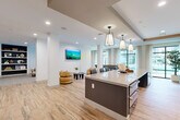 Located in Houston's vibrant 77007 corridor, this single-floor amenity lounge offers a thoughtfully designed open-plan space that seamlessly blends social and relaxation zones. The layout is anchored by a large central island topped with a crisp quartz surface and flanked by rich wood-toned cabinetry, illuminated by a pair of geometric pendant lights. A dedicated café-style bar area features a bold hexagonal tile backsplash in warm copper and charcoal tones, a coffee station, and bar seating with leather-accented chairs — ideal for casual gatherings or focused work. Light wood-look flooring runs throughout, lending warmth and continuity across the space. A lounge area transitions seamlessly into a carpeted sitting room furnished with neutral sofas, accent chairs, and built-in shelving with dark contrasting backing, creating an inviting retreat. Floor-to-ceiling windows along one wall bathe the interior in natural light while offering outdoor views. The entry corridor features a textured wood-grain accent wall and a striking arched iron-framed mirror, setting an elegant tone. Recessed lighting, wall-mounted displays, and carefully curated décor complete the polished, contemporary aesthetic of this well-appointed communal space.