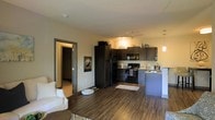 This single-floor residence offers a thoughtfully designed open-concept layout that seamlessly connects the living area to the kitchen, creating a spacious and functional flow. The living area features rich hardwood-style flooring and is comfortably appointed, with a bright, neutral color palette that lends a warm and welcoming ambiance throughout.

The kitchen is well-equipped with dark, espresso-toned cabinetry that provides ample storage, black appliances including a refrigerator, range, and microwave, and a light-toned countertop with a sink overlooking the space. A pendant light fixture adds a stylish accent above the kitchen area, complementing the overall modern aesthetic.

A doorway off the main living space leads to an adjacent room with carpeted flooring, offering a comfortable contrast to the hardwood in the main area. The transition between spaces is framed by clean architectural detailing, including crisp door trim and smooth ceilings.

Natural and artificial lighting work in harmony to keep the interior bright and inviting. The overall design balances functionality with contemporary style, making this a practical and attractive single-level living space suited for everyday comfort.