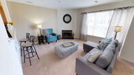 This single-floor model unit showcases a thoughtfully designed open-concept layout that connects the living area and kitchen in a seamless, functional arrangement. The living room is generously proportioned, featuring plush wall-to-wall carpeting in a neutral tone that complements the warm beige walls and white ceiling. A large window dressed with patterned grommet curtains allows natural light to fill the space, creating a bright and airy atmosphere. A freestanding fireplace serves as a focal point along one wall, adding both warmth and visual interest to the room. The living area comfortably accommodates a sectional sofa, accent chair, and an upholstered ottoman, illustrating the versatility of the floor plan.

The kitchen is accessible via a pass-through counter with bar seating, making it ideal for casual dining and entertaining. Warm oak cabinetry lines the kitchen walls, paired with standard appliances and overhead lighting. Decorative wall accents near the pass-through add a personalized, homey touch. Overall, the unit presents a practical and inviting single-level living environment with a cohesive design aesthetic throughout.