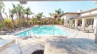 This outdoor amenity space exudes a resort-like atmosphere, centered around a generously sized, crystal-clear swimming pool surrounded by a spacious tiled deck. The pool area is well-appointed with rows of lounge chairs positioned to capture plenty of sunshine, and shade umbrellas offer a respite from the sun on warm days. Towering palm trees and lush tropical landscaping frame the perimeter, creating a vibrant, vacation-inspired ambiance. The surrounding architecture features a warm Mediterranean-inspired aesthetic, with stucco exteriors, terracotta-tiled rooflines, and warm earth tones that complement the outdoor setting. A covered pavilion serves as the centerpiece for outdoor entertaining, equipped with a built-in stainless steel gas grill and generous granite-topped countertops. The wood-beamed ceiling overhead provides ample shade, while the open sides allow for natural airflow and views of the greenery beyond. Tiled accent walls and premium granite surfaces throughout the BBQ area reflect a high level of finish quality. The combination of a resort-style pool, professional-grade outdoor cooking facilities, and lush landscaping makes this a standout space designed for both leisure and social gatherings.