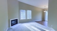 This single-floor residence showcases a thoughtfully designed living space with standout architectural details. The main living area draws immediate attention with its soaring vaulted ceilings, which lend an airy, open quality to the room. Soft, neutral green walls create a calming atmosphere throughout, complemented by plush wall-to-wall carpeting that adds warmth underfoot, while adjacent areas transition to smooth hardwood-style flooring. A classic fireplace with a clean white surround and dark insert serves as a natural focal point, making the living room an inviting space for relaxation. Two generously sized windows flood the room with abundant natural light, casting bright, sweeping rays across the interior. These windows also frame views of lush, verdant greenery outside, seamlessly connecting the interior to the natural surroundings. A pendant light fixture is partially visible, hinting at thoughtful lighting design throughout the home. The overall ambiance of this space is bright, open, and welcoming, with a clean and move-in-ready presentation that offers a versatile canvas for a variety of interior styles.