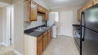 This single-floor unit offers a practical and efficiently arranged layout across two bedrooms and one bathroom. Light beige tile flooring runs consistently throughout the entire space, creating a cohesive and easy-to-maintain interior. Neutral-toned walls in a warm tan hue complement the flooring and contribute to a bright, open atmosphere.

The open living area connects directly to the kitchen, which is equipped with warm oak cabinetry, dark laminate countertops, a double-basin sink, and black appliances including a refrigerator and range. A notable feature of the kitchen is the inclusion of an in-unit stacked washer and dryer, along with a door providing exterior access.

Both bedrooms are generously proportioned for the overall footprint of the unit, each featuring windows with exterior views that allow natural light to filter in throughout the day. The rooms share a clean, simple design with the same neutral wall tones and tile flooring found throughout the rest of the space.

The single bathroom includes a white vanity with under-cabinet storage, a toilet, and a tub-shower combination enclosed by a curtain. Tile detailing on the lower walls adds a functional and finished touch. Ceiling-mounted light fixtures provide consistent illumination across all rooms.