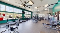 This fitness center in Lakewood, Colorado offers a comprehensive workout environment designed to support a full range of exercise routines. The space is defined by its high ceilings and expansive floor-to-ceiling windows along one wall, which flood the interior with abundant natural light and provide open views of the outdoor pool area and surrounding greenery — an energizing backdrop for any workout.

The interior design features bold teal and green accent walls set against light gray tones, creating a vibrant yet focused atmosphere. Durable rubber flooring spans the entire space, providing both safety and functionality. Large ceiling fans contribute to air circulation throughout.

The cardio zone is well-appointed with multiple treadmills and stationary bikes positioned directly in front of the large windows. A wall-mounted flat-screen television is also available in this area. The strength training section offers a diverse selection of equipment, including cable machines, a multi-function rack with suspension training accessories, selectorized weight machines, and a dedicated free weights area featuring a full rack of dumbbells and adjustable benches. Floor mirrors line one wall, aiding in form and expanding the sense of space. Recessed and panel lighting throughout ensures consistent illumination across all workout zones.