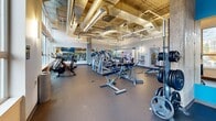 Located at 337 NE 103rd St in Seattle, WA, this fitness center offers a spacious, single-floor open layout designed to accommodate a comprehensive range of workout activities. The space features an industrial-modern aesthetic, highlighted by exposed concrete columns, high ceilings with visible ductwork, and polished metal ventilation systems that lend a raw yet refined character to the environment.

Large floor-to-ceiling windows line the perimeter walls, flooding the interior with abundant natural light and offering views of the surrounding greenery. The durable rubber flooring throughout provides both safety and functionality, supporting a wide variety of training activities.

The cardio zone is well-stocked with treadmills, elliptical machines, and stationary bikes, all arranged to take advantage of the natural light and outdoor views. The strength training area features a robust selection of free weights, kettlebells, barbells, and adjustable benches, alongside cable and resistance machines. An accent wall featuring motivational fitness graphics in bold green adds an energetic visual element to the space.

Full-length mirrors are strategically placed to assist with form and to enhance the sense of openness. The layout is thoughtfully organized to allow smooth circulation between zones, making this fitness center a functional and inviting space for all levels of fitness enthusiasts.
