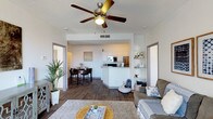 This single-floor residence offers an open-concept layout that thoughtfully connects the living, dining, and kitchen areas into one cohesive, functional space. The living room is anchored by a ceiling fan with a light fixture and features rich, dark hardwood-style flooring that extends throughout the main living areas. A plush sofa, woven coffee table, and layered area rug create a warm and inviting atmosphere, complemented by built-in shelving along one wall — ideal for displaying décor and personal items.

The dining area sits just off the living space and comfortably accommodates a table and bar-style seating, making it perfect for both casual meals and entertaining. A lush floor plant adds a touch of natural greenery to the corner, enhancing the overall warmth of the room.

The kitchen features white cabinetry, a breakfast bar peninsula, and stainless steel appliances, including a refrigerator and microwave. The white-on-white palette keeps the kitchen feeling bright and clean, while the open pass-through design maintains the airy, connected feel of the space. The textured ceiling and neutral white walls throughout provide a clean backdrop that ties each area together cohesively.