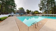 Located at 201 Treybrooke Dr in Morrisville, North Carolina, this community pool facility offers a well-appointed outdoor aquatic space set against a backdrop of towering pine trees. The centerpiece is a generously sized swimming pool with crystal-clear blue water, accessible via stainless steel entry rails and surrounded by an expansive concrete deck. The deck is furnished with multiple lounge chairs and shaded seating areas featuring blue umbrellas, providing ample space for relaxation and recreation. A blue-sided clubhouse structure anchors one side of the facility, featuring a covered upper-level porch with large windows overlooking the pool area. The entire facility is enclosed by a classic white vinyl fence, which is complemented by neatly trimmed hedgerows and landscaped garden beds. A dedicated entry point with gating and posted pool rules ensures orderly access. The natural canopy of mature pine trees surrounding the perimeter provides privacy and shade, enhancing the serene outdoor ambiance. Additional amenities include conveniently placed waste receptacles and practical utility access points, making this a fully functional and well-maintained community gathering space.
