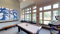 The game room is a well-appointed recreational space designed for both entertainment and comfort. The room's most striking architectural feature is its dramatic two-story wall of large, grid-paned windows that flood the interior with abundant natural light while offering expansive views of the surrounding greenery and landscaping. The soaring ceiling height enhances the open, airy feel of the space, making it feel generous and inviting. A stylish chandelier adds a refined touch overhead. The centerpiece of the room is a full-size billiards table with a classic dark wood frame and cream felt surface, fully racked and ready for play. Neutral-toned carpeting runs underfoot, providing a comfortable surface throughout. A bold blue abstract artwork is displayed prominently on one wall, adding a contemporary artistic element that complements the modern design aesthetic. Sleek wall sconces provide additional ambient lighting. A low-profile bench runs along the base of the window wall, offering casual seating. The overall ambiance is modern and welcoming, balancing a clean architectural design with recreational functionality.