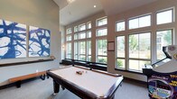 The game room is a well-appointed recreational space designed for both entertainment and comfort. The room's most striking architectural feature is its dramatic two-story wall of large, grid-paned windows that flood the interior with abundant natural light while offering expansive views of the surrounding greenery and landscaping. The soaring ceiling height enhances the open, airy feel of the space, making it feel generous and inviting. A stylish chandelier adds a refined touch overhead. The centerpiece of the room is a full-size billiards table with a classic dark wood frame and cream felt surface, fully racked and ready for play. Neutral-toned carpeting runs underfoot, providing a comfortable surface throughout. A bold blue abstract artwork is displayed prominently on one wall, adding a contemporary artistic element that complements the modern design aesthetic. Sleek wall sconces provide additional ambient lighting. A low-profile bench runs along the base of the window wall, offering casual seating. The overall ambiance is modern and welcoming, balancing a clean architectural design with recreational functionality.