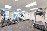 Located at 1477 Mississauga Valley Blvd in Mississauga, Ontario, this well-equipped fitness centre offers a comprehensive workout facility across a single floor. The space is divided into distinct functional zones, providing users with a versatile exercise environment. The cardio area features treadmills, elliptical trainers, and a stair climber, all positioned near large windows with vertical blinds that allow natural light to fill the room. A wall-mounted flat-screen television adds convenience for those using the cardio equipment. The strength training section includes a multi-functional cable machine, a free weights bench, and a barbell, complemented by large wall-mounted mirrors that enhance the sense of space and allow users to monitor their form. A dedicated stretching and functional training area offers a padded adjustable bench and additional open floor space, with a stretching guide poster displayed on the wall for reference. Throughout the facility, recessed panel lighting in the textured ceiling ensures bright, even illumination. The flooring transitions between durable carpet tiles in the cardio zone and rubber-style flooring in the strength area, each suited to its respective use. A wall clock and posted workout guides contribute to the practical, informative atmosphere of this fully functional fitness space.