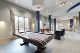 This single-floor amenity space is designed with a contemporary aesthetic, characterized by a striking navy blue and warm grey color palette that carries consistently throughout. The open layout connects multiple functional zones seamlessly, creating a cohesive and inviting environment.

The entry area features light-toned large-format tile flooring, curved architectural walls painted in deep navy blue, and decorative metallic wall art, setting an immediately polished tone. Adjacent seating lounges offer comfortable upholstered sofas and armchairs, sconce lighting, and natural light from large windows with patterned drapery.

The central hub features a curved granite-topped island with mosaic tile detailing and pendant lighting overhead, flanked by navy-painted structural columns. A fully equipped kitchen nearby includes rich wood cabinetry, stainless steel appliances, a mosaic tile backsplash, and a granite breakfast bar with seating.

A dedicated workstation area provides high-top tables and modern seating, while a separate game room features a full-sized billiards table, ceiling fans, and generous natural light from floor-to-ceiling windows. Throughout the space, recessed lighting, bold accent walls, and carefully curated furnishings contribute to a refined yet energetic community atmosphere.