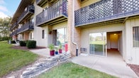 This single-floor unit offers a practical and comfortable layout designed for easy living. The exterior presents a classic multi-unit building aesthetic with brick and vertical siding accents, decorative lattice-railed balconies above, and a private ground-level concrete patio accessible via sliding glass doors — a charming outdoor space with room for potted plants and relaxation.

Inside, the main living area is open and generously proportioned, featuring neutral wall-to-wall carpeting, crisp white walls, and a ceiling fan that adds both function and a touch of warmth to the space. Baseboard heating runs along the perimeter, and a wall-mounted air conditioning unit ensures year-round comfort. The open layout flows naturally toward the kitchen, which is visible through a wide doorway and is equipped with a gas range and refrigerator, as well as tile flooring that contrasts with the carpeted living areas.

Additional rooms branch off from the main living space, providing flexible options for sleeping or storage. Sliding glass doors along one wall bathe the interior in natural light while framing views of the well-maintained lawn and the lush greenery beyond. The overall ambiance is clean, bright, and move-in ready, offering a comfortable retreat within a well-established residential community.