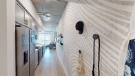 Located at 201 The Promenade N in Long Beach, California, this single-floor urban loft showcases an industrial-chic aesthetic defined by exposed concrete ceilings with visible ductwork and structural columns throughout. The layout flows from a well-appointed entry hallway, featuring a striking swirling patterned accent wall, into the combined kitchen and living area at its core.

The kitchen is sleek and functional, outfitted with gray shaker cabinetry, white quartz countertops, stainless steel appliances including a gas range and refrigerator, and a wine cooler with brass hardware accents. Pendant lighting in black and gold hangs above the countertop, adding warmth to the space. Light wood-tone flooring runs throughout, complementing the neutral palette.

The living area extends from the kitchen, offering an open, airy feel with natural light filtering in from a window at the far end. Exposed concrete columns punctuate the space, reinforcing the loft's industrial character.

The bedroom features a bold geometric accent wall, an exposed concrete ceiling, and direct access to the bathroom. The bathroom is clean and modern, with tile surround, a soaking tub with shower, built-in shelving, and a white vanity. A dedicated closet completes the layout.