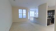 This single-floor space features an open-concept layout that seamlessly connects the living area to the kitchen. The living area is finished with neutral beige carpet and warm-toned walls, creating a clean and inviting atmosphere. Multiple windows fitted with horizontal blinds allow generous natural light to fill the space throughout the day.

The kitchen is defined by a curved peninsula counter that serves as a natural divider between the cooking and living zones. Overhead, dual pendant lights hang above the peninsula, adding focused accent lighting and a subtle decorative touch. A track lighting fixture is also installed on the kitchen ceiling, providing additional illumination for the workspace. The peninsula includes an integrated sink, making it both functional and visually central to the space.

Beyond the kitchen, a secondary windowed area is visible, suggesting additional living or dining space that benefits from further natural light. The overall design is neutral and understated, with consistent finishes throughout that contribute to a cohesive, well-lit, and functional living environment.