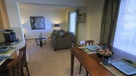 This single-floor space features an open-concept layout that seamlessly integrates the dining area and living room into one cohesive environment. The carpeted flooring in neutral beige tones extends throughout, contributing to a warm and inviting atmosphere. The living area is anchored by a plush, dark upholstered sofa accented with patterned throw pillows, accompanied by a sleek glass-top coffee table. A framed piece of wall art adds a decorative focal point to the living space. A window unit air conditioner is installed near a window draped with light curtains, allowing natural light to filter into the room. The dining area, positioned in the foreground, features a rich, dark wood table paired with matching wooden chairs, providing a functional and stylish space for meals. Green curtain panels frame the right side of the space, adding a subtle pop of color against the neutral walls. The open layout creates a sense of generous space, making the area feel connected and easy to navigate. Overall, the space presents a practical and comfortable design suitable for everyday living.