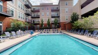 Located at 55 E 9th Ave in Columbus, Ohio, this outdoor amenity space centers around a generously sized swimming pool that serves as the focal point of the community's courtyard. The pool features clean, clear blue water and is framed by a wide pool deck lined with rows of lounge chairs and seating, providing ample space for residents to relax and unwind. The courtyard is enclosed by the surrounding multi-story residential building, which features a combination of brick and neutral-toned siding, along with private balconies that overlook the pool area. Young trees are thoughtfully planted throughout the space, adding greenery and a welcoming natural element to the hardscape environment. A structured parking facility is visible adjacent to the pool area, indicating convenient on-site parking for residents. The open-air design of the courtyard allows for abundant natural light, making it an inviting setting during warmer months. The overall ambiance is that of a well-appointed, community-oriented outdoor retreat, designed to encourage social interaction and leisure within an urban residential setting in Columbus.