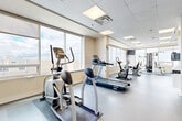Located at 725 Highway 15 in Kingston, Ontario, this fitness facility offers a generously sized, single-floor exercise room designed to support a comprehensive workout experience. The space is finished with durable flooring throughout and features a drop-panel ceiling outfitted with recessed panel lighting that ensures bright, even illumination across the entire area.

Large windows line the perimeter walls, flooding the room with natural light and providing open views of the surrounding skyline — an invigorating backdrop for any workout. The neutral, warm-white wall tones and clean architectural lines create a bright, welcoming ambiance.

The exercise room is thoughtfully laid out to accommodate a wide variety of fitness equipment, including treadmills, elliptical trainers, upright and recumbent stationary bikes, a multi-station cable and weight machine, a rowing machine, and a stability ball station. Wall-mounted flat-screen televisions are positioned throughout the space for entertainment during workouts. Structural columns divide portions of the floor plan, creating natural zones within the open layout.

Additional features include a wall-mounted clock, motivational signage, and dedicated entry doors for convenient access. The spacious, open floor plan provides ample room to move freely between equipment stations, making this facility suitable for a range of fitness activities and user needs.