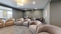 Located in Centennial, Colorado, this well-appointed community amenity floor offers a variety of distinct gathering spaces designed to encourage both social interaction and quiet relaxation. The layout is organized and flowing, with a hallway connecting the key zones throughout.

The central dining and lounge area is a visual highlight, featuring a coffered ceiling, warm wood built-in shelving lined with books and décor, and a large dining table surrounded by teal upholstered chairs. Adjacent to this is a teal channel-tufted banquette seating nook, accented by warm wall sconces, creating an intimate dining atmosphere.

A broader open lounge area offers flexible seating with plush rounded armchairs and mobile lounge chairs, ideal for casual gatherings. Natural light enters through large windows, and a black metal stair railing defines the open upper-level overlook. A dedicated media room features a wall-mounted flat-screen television, soft seating, and a cozy, cinema-inspired setup.

The kitchen provides a service-oriented counter space visible from the main lounge. Throughout, neutral-toned carpet, sage green walls, and recessed lighting create a calm, modern ambiance that balances comfort with contemporary design.