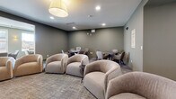 Located in Centennial, Colorado, this well-appointed community amenity floor offers a variety of distinct gathering spaces designed to encourage both social interaction and quiet relaxation. The layout is organized and flowing, with a hallway connecting the key zones throughout.

The central dining and lounge area is a visual highlight, featuring a coffered ceiling, warm wood built-in shelving lined with books and décor, and a large dining table surrounded by teal upholstered chairs. Adjacent to this is a teal channel-tufted banquette seating nook, accented by warm wall sconces, creating an intimate dining atmosphere.

A broader open lounge area offers flexible seating with plush rounded armchairs and mobile lounge chairs, ideal for casual gatherings. Natural light enters through large windows, and a black metal stair railing defines the open upper-level overlook. A dedicated media room features a wall-mounted flat-screen television, soft seating, and a cozy, cinema-inspired setup.

The kitchen provides a service-oriented counter space visible from the main lounge. Throughout, neutral-toned carpet, sage green walls, and recessed lighting create a calm, modern ambiance that balances comfort with contemporary design.