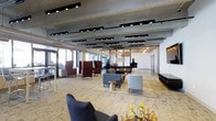 This contemporary amenity space is thoughtfully designed across a single level, combining an expansive interior lounge with a covered outdoor terrace. The interior lounge showcases an industrial-chic aesthetic, highlighted by exposed concrete ceilings with visible ductwork, bold track lighting, and wide windows that flood the space with natural light. Eclectic seating arrangements — including statement wingback chairs in deep burgundy, plush sofas, and accent side tables — are grouped across patterned area rugs, creating inviting conversation zones. A reception or service desk anchors the far end of the room, lending the space a polished, hospitality-inspired feel. The covered outdoor terrace offers a seamlessly connected extension of the interior, featuring modular gray sectional sofas, slatted wood accent tables, and additional lounge chairs arranged for relaxed gathering. Recessed lighting illuminates the paneled ceiling overhead, while large structural columns frame open views of the surrounding urban streetscape and neighboring buildings. Lush potted plants and planters with tall ornamental grasses add greenery throughout, softening the modern palette. A grilling station and game table area enhance the terrace's functionality, making it well-suited for both casual relaxation and social entertaining.