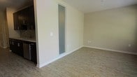 This single-floor space offers a straightforward and functional layout with a clean, contemporary aesthetic. The living area is finished with light-toned hardwood-style flooring that runs throughout, lending warmth and continuity to the space. Neutral beige walls provide a versatile backdrop, complemented by crisp white baseboards that frame the room neatly.

A standout architectural detail is the slim, frosted glass panel inset into the interior wall, which introduces a modern design element while allowing diffused light to pass between spaces without sacrificing privacy. This feature adds visual interest to an otherwise understated interior.

Adjacent to the main living area, a partial view of what appears to be a kitchen is visible, featuring dark cabinetry that contrasts with the lighter tones of the living space. Standard electrical outlets are positioned along the walls, and a light switch is conveniently placed near the glass panel.

The overall ambiance is minimal and move-in ready, with a neutral palette that offers flexibility for personalization. The open feel of the main living area, combined with the practical finishes, makes this a functional and adaptable space suitable for a variety of uses.