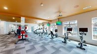 Located at 5280 Beechmont Ave in Cincinnati, OH, this single-floor fitness center is thoughtfully designed to support a full range of workout activities. The space is defined by its striking bold orange ceilings, crisp white walls, and patterned carpet tile flooring throughout the main exercise areas, creating an energetic yet clean aesthetic.

The primary cardio floor is generously proportioned, accommodating a wide array of machines including treadmills, ellipticals, stair climbers, and stationary bikes—each equipped with individual screens for entertainment. Multiple ceiling-mounted flat-screen TVs and ceiling fans are installed throughout, ensuring a comfortable environment. Large windows along the perimeter flood the space with natural light and offer views of the adjacent outdoor area.

A dedicated group fitness and cycling studio features light hardwood flooring, floor-to-ceiling mirrors, large windows, and a ceiling fan, providing an inviting space for spin classes and group workouts. An adjacent functional training area offers hardwood flooring, a full wall of mirrors, and a well-stocked equipment rack with stability balls, resistance tools, and step platforms.

The free weight section includes a dumbbell rack, weight bench, and cable machine, supported by mirrored walls. A separate recovery or stretching room with carpeted flooring, a padded bench, cabinetry, and a ceiling fan rounds out the facility, offering a calm space for post-workout recovery.