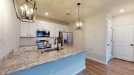 Located at 6480 Alexander Clay in Columbus, Ohio, this well-appointed single-floor home offers a thoughtfully designed open-concept layout. The kitchen is a standout feature, showcasing crisp white shaker cabinetry, granite countertops, a decorative tile backsplash, and a full suite of stainless steel appliances including a French door refrigerator, electric range, microwave, and dishwasher. A large central island with an undermount sink and matte black faucet serves as both a prep space and gathering spot, illuminated by elegant geometric cage pendant lights. The open living and dining areas flow seamlessly from the kitchen, featuring warm hardwood-style flooring and generous windows that invite abundant natural light. Carpeted rooms offer a comfortable retreat, with exterior door access to an outdoor area. The primary bathroom features a granite-topped double vanity with white cabinetry, matte black hardware, a glass-enclosed shower, and direct access to a walk-in closet with built-in shelving. A dedicated laundry area houses a stacked washer and dryer unit for added convenience. Neutral tones, clean lines, and quality finishes throughout give this home a modern yet inviting character.