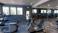 This single-floor fitness center offers a thoughtfully designed workout space that combines functionality with a modern aesthetic. The interior is defined by deep teal-painted walls that create an energizing atmosphere, complemented by recessed lighting and a textured ceiling that adds depth to the space. Large windows along the walls allow natural light to fill the room while providing views of the surrounding greenery outdoors, creating an open and inviting environment. The flooring features a distinctive multi-toned carpet tile pattern in shades of blue, gray, and tan, adding a dynamic visual element underfoot. The facility is outfitted with a range of cardio equipment, including stationary bikes and an elliptical trainer from Precor, as well as a multi-station strength training machine positioned near a large wall mirror that visually expands the space and allows users to monitor their form. Storage shelving and equipment racks are neatly arranged along the walls, keeping the area organized and accessible. A glass-panel door provides direct access to the exterior, further enhancing the sense of openness. Overall, the fitness center delivers a clean, well-maintained environment suitable for a full range of workout routines.