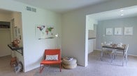 This single-story residence at TGM Autumn Woods offers a clean, open layout designed for comfortable everyday living. The living area features neutral wall tones and soft carpeting underfoot, creating a welcoming atmosphere that feels both bright and understated. A colorful world map print adds a lively focal point to the room, while the space itself is versatile enough to accommodate a variety of furniture arrangements. A half-wall partition provides a natural visual separation between the living area and the adjacent kitchen without closing off the floor plan, maintaining an open, airy feel throughout. The kitchen, visible through the passthrough opening, features dark countertops that contrast with the lighter cabinetry and walls. Just beyond, a dining area with a glass-top table and modern chairs completes the connected living zone. Natural light filters through the space, contributing to its bright and neutral ambiance. The overall design emphasizes simplicity and functionality, with consistent finishes throughout that give the home a cohesive, well-maintained appearance. The single-floor layout ensures easy accessibility and a straightforward flow between all areas of the residence.