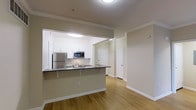 This single-floor residence is thoughtfully designed with an open-concept layout that seamlessly connects the kitchen and living areas, creating a bright and functional living environment. The kitchen is a standout feature, boasting crisp white cabinetry that extends to the ceiling, complemented by dark countertops that provide a striking contrast. A peninsula with bar-top seating serves as both a practical workspace and a casual dining area. Stainless steel appliances, including a full-size refrigerator and a built-in microwave, add a sleek, modern touch. Crown molding runs throughout the space, lending an elevated, refined character to the interiors. Warm hardwood-style flooring flows continuously across the main living area, contributing to a cohesive and inviting atmosphere. The neutral, earthy wall tones provide a versatile backdrop suitable for a variety of décor styles. A corridor visible from the main living space leads to additional rooms, each framed by clean, paneled white doors. The overall ambiance is clean, contemporary, and well-appointed, making this a comfortable and stylish single-level residence.